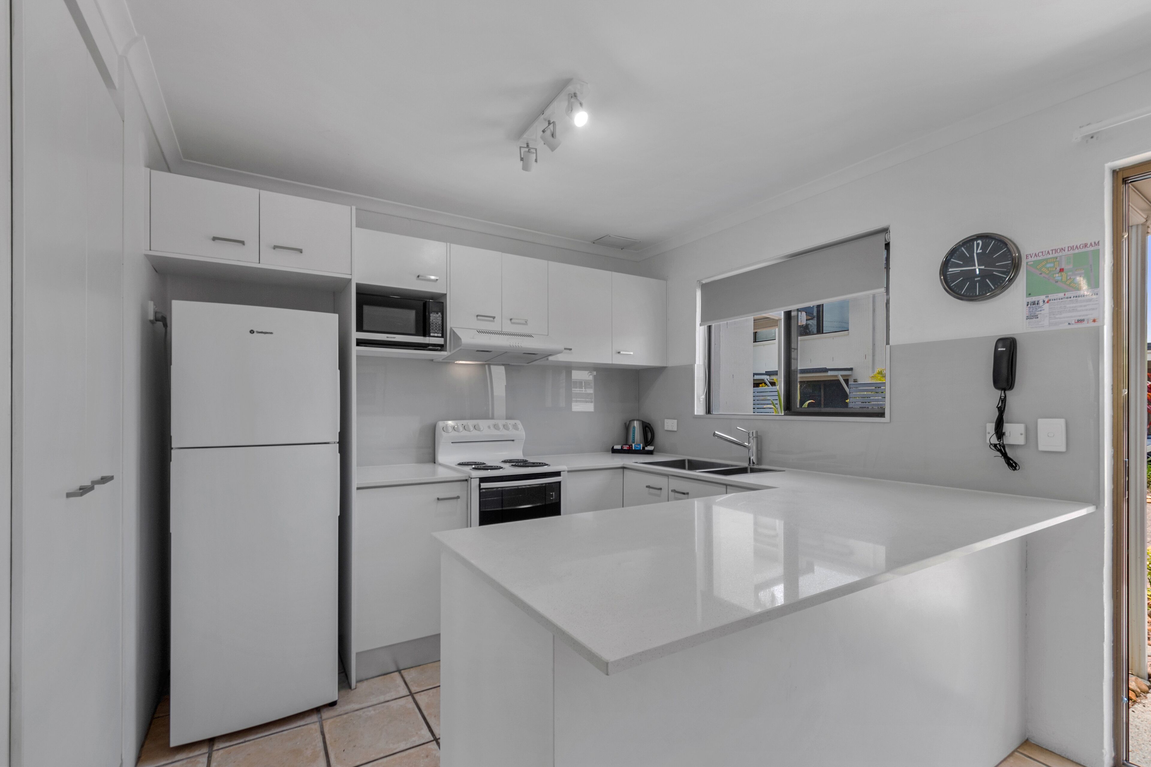 A modern kitchen with white cabinetry, a built-in microwave, a stove, and a sink. There is a clock on the wall and an evacuation diagram.