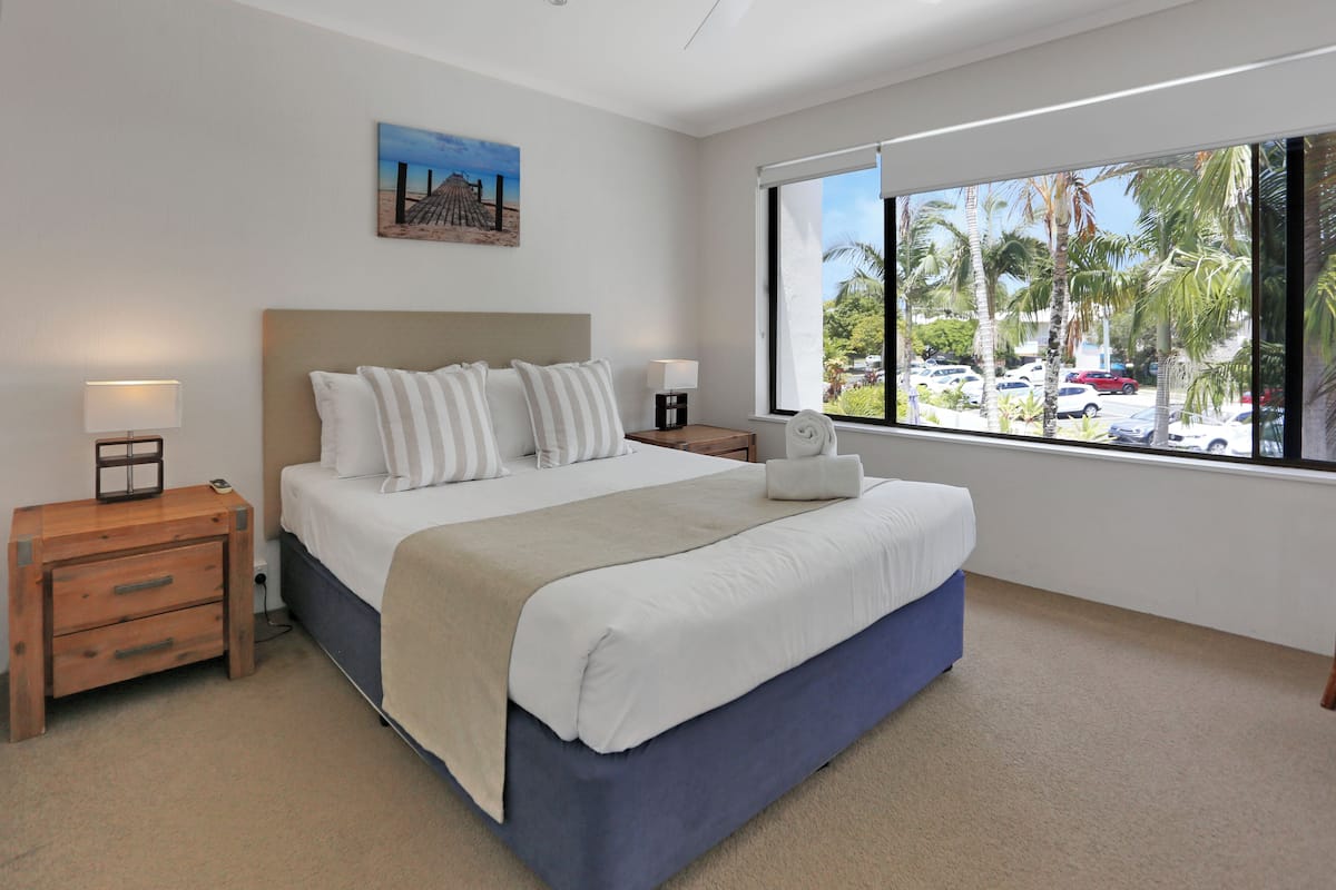 A bedroom with a bed, bedside tables, a window with a view of parked cars and palm trees, and a painting on the wall.