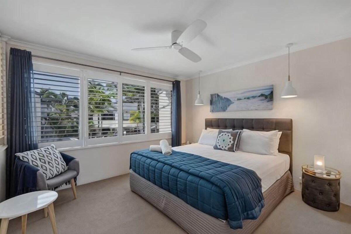 A bedroom with a bed, a chair, a table, a ceiling fan, and a window with blinds.