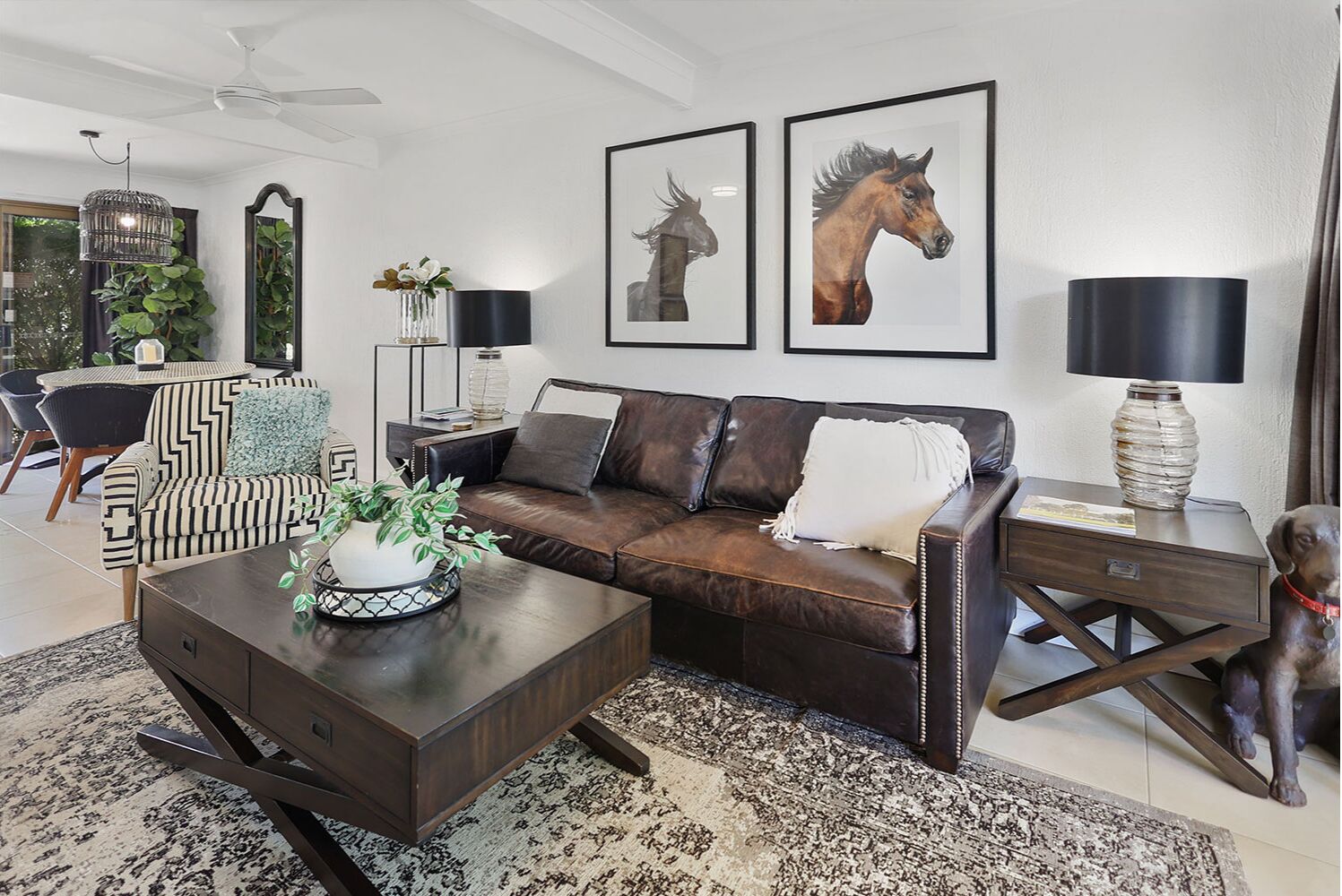 A modern living room with a dark brown leather sofa, a striped armchair, a coffee table with a plant, and framed horse paintings on the wall.