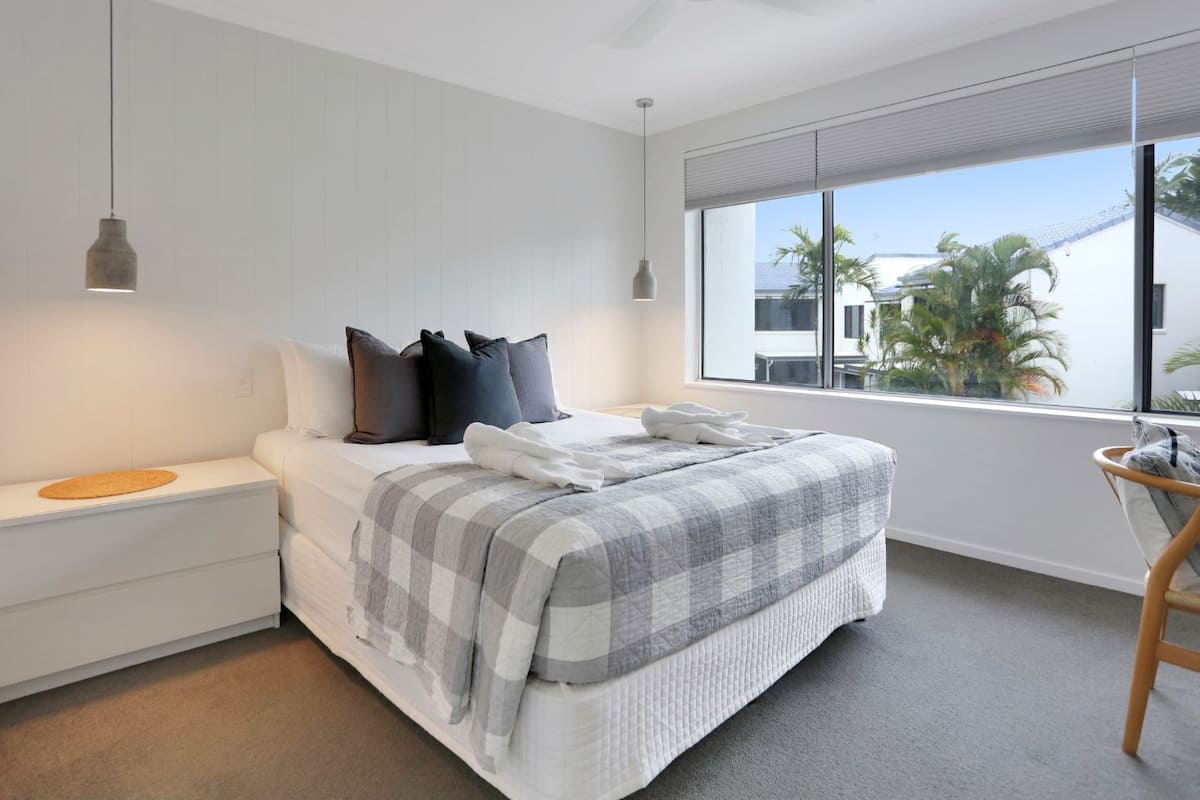 A modern bedroom with a bed, bedside tables, a window with a view of palm trees, and a ceiling fan.