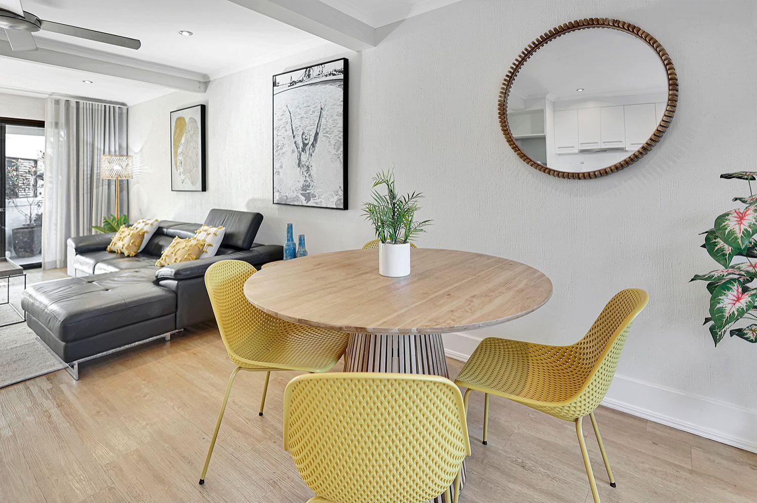 A modern living room with a round wooden dining table, yellow woven chairs, a grey sofa with decorative pillows, a large mirror, and a potted plant.