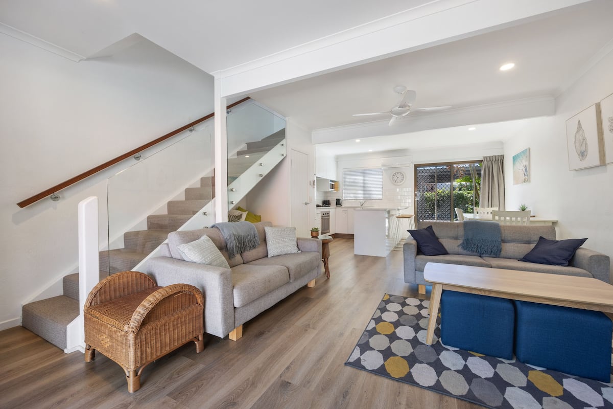 A spacious living area with a staircase, two sofas, a dining table, and a kitchen in the background.