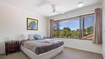 A bedroom with a large bed, a nightstand, a ceiling fan, and a window with a view of trees and buildings.