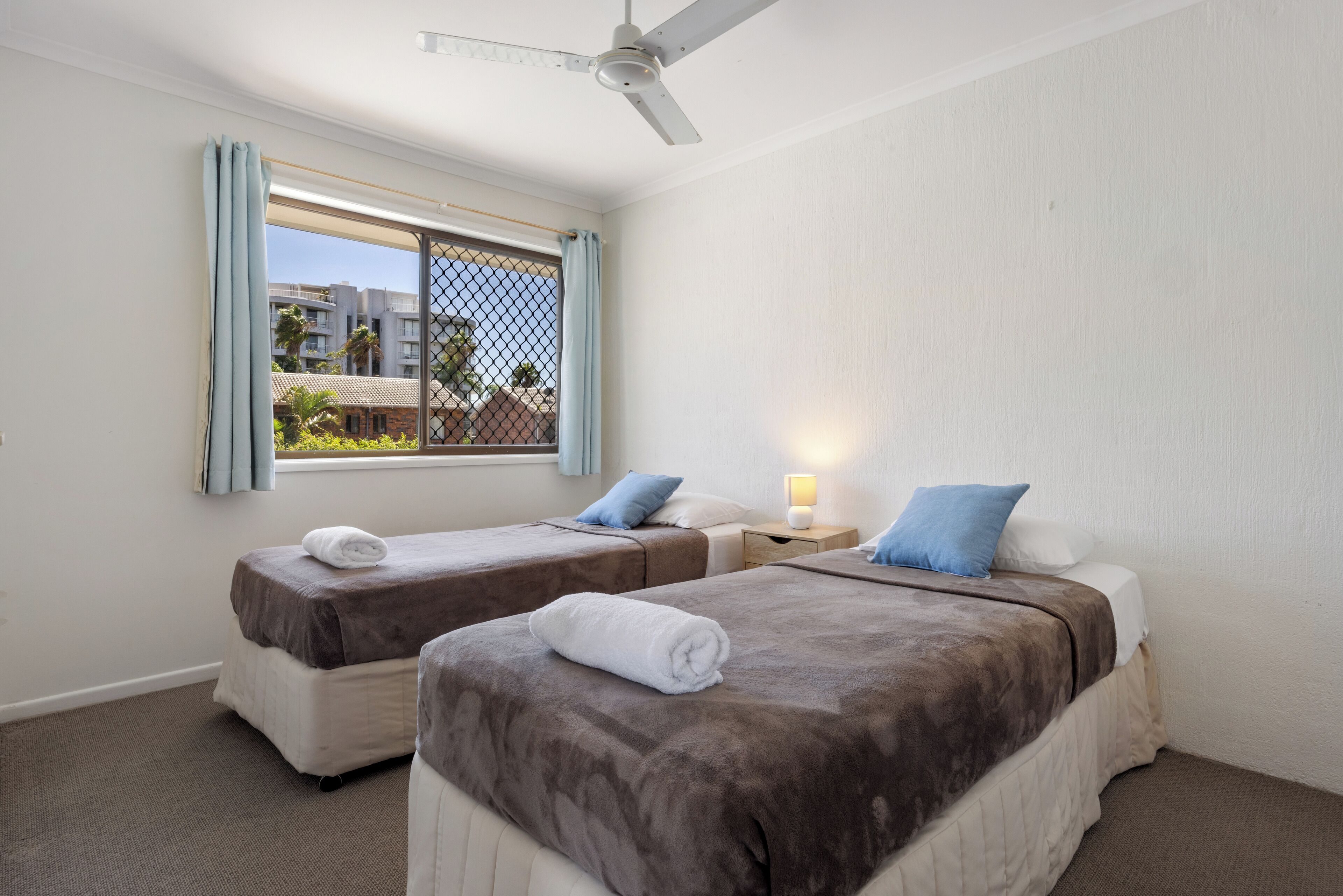 Une chambre d’hôtel avec deux lits, un ventilateur de plafond, une fenêtre avec des rideaux et une vue sur les bâtiments et la verdure environnante.