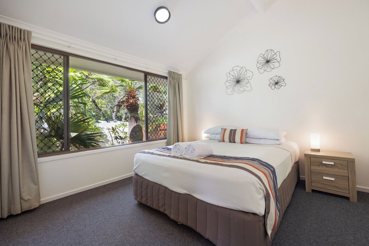 A hotel room with a large window, a bed with a striped blanket, a bedside table with a lamp, and wall art.