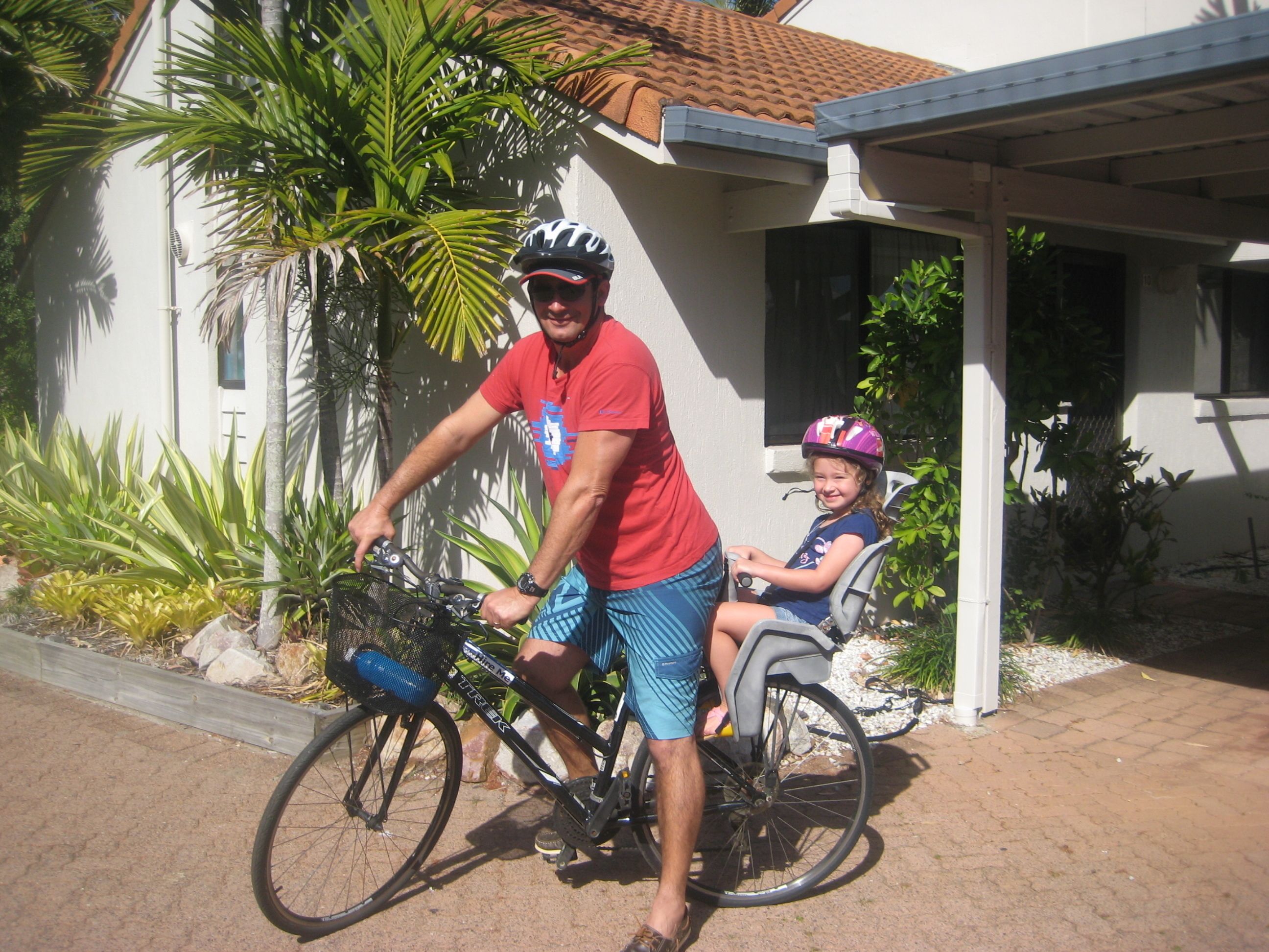 A man and a child on a bicycle in front of a house with a tiled roof and a porch.