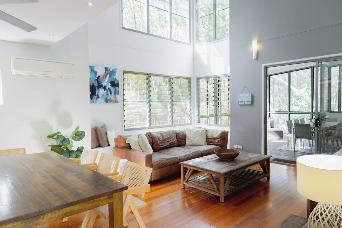 A spacious living area with a wooden dining table, a sofa, and a coffee table. There are large windows providing natural light and a view of the outdoors.