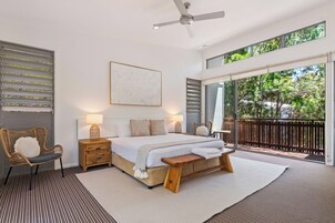 A bedroom with a large bed, a bedside table, a chair, a ceiling fan, and a view of the outdoors through a sliding glass door.