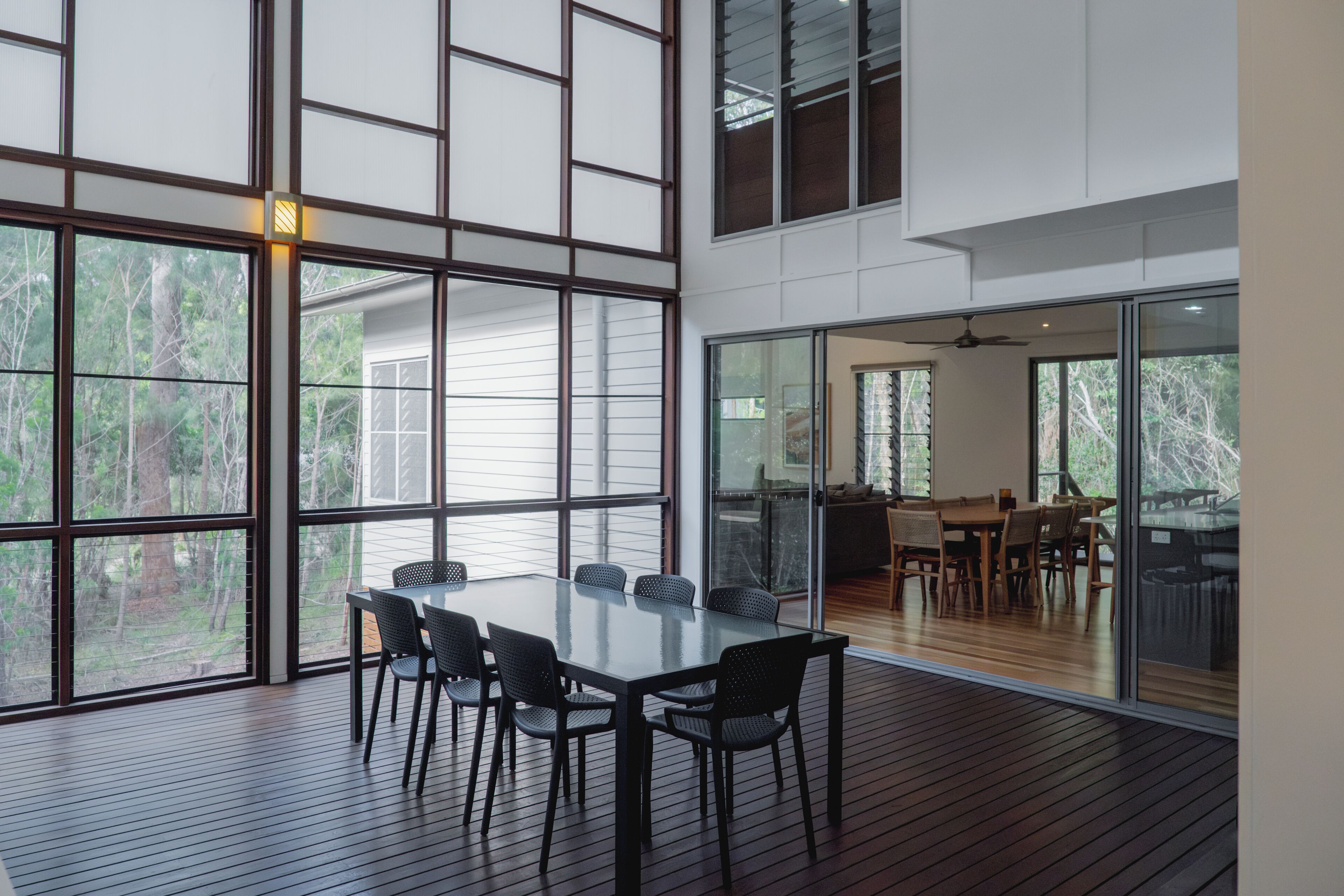 Een modern interieur met een grote eettafel en stoelen, grote ramen met uitzicht op bomen, en een glazen scheidingswand die het eetgedeelte van een andere ruimte afscheidt.