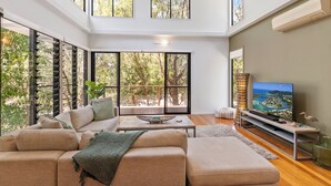 A modern living room with a large sectional sofa, a flat-screen TV, and large windows offering a view of trees and a deck.