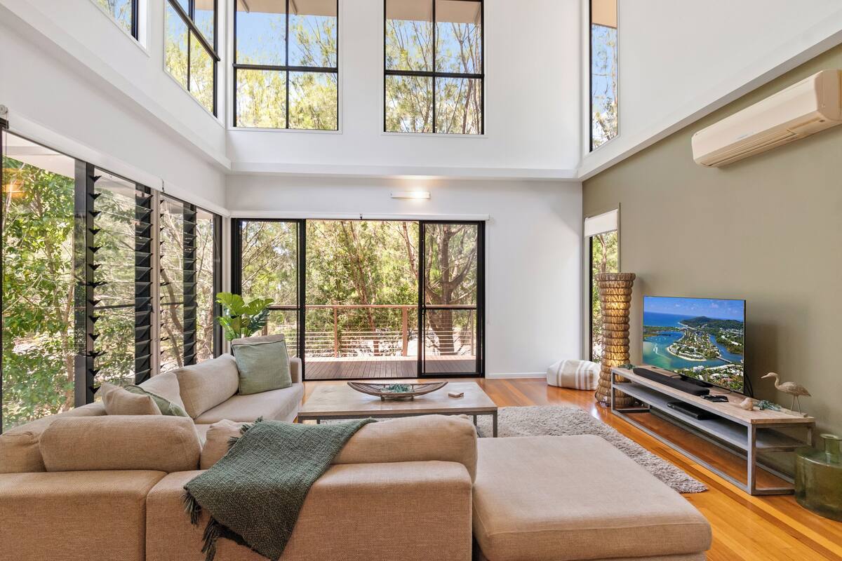 A modern living room with a large sectional sofa, a flat-screen TV, and large windows offering a view of trees and a deck.