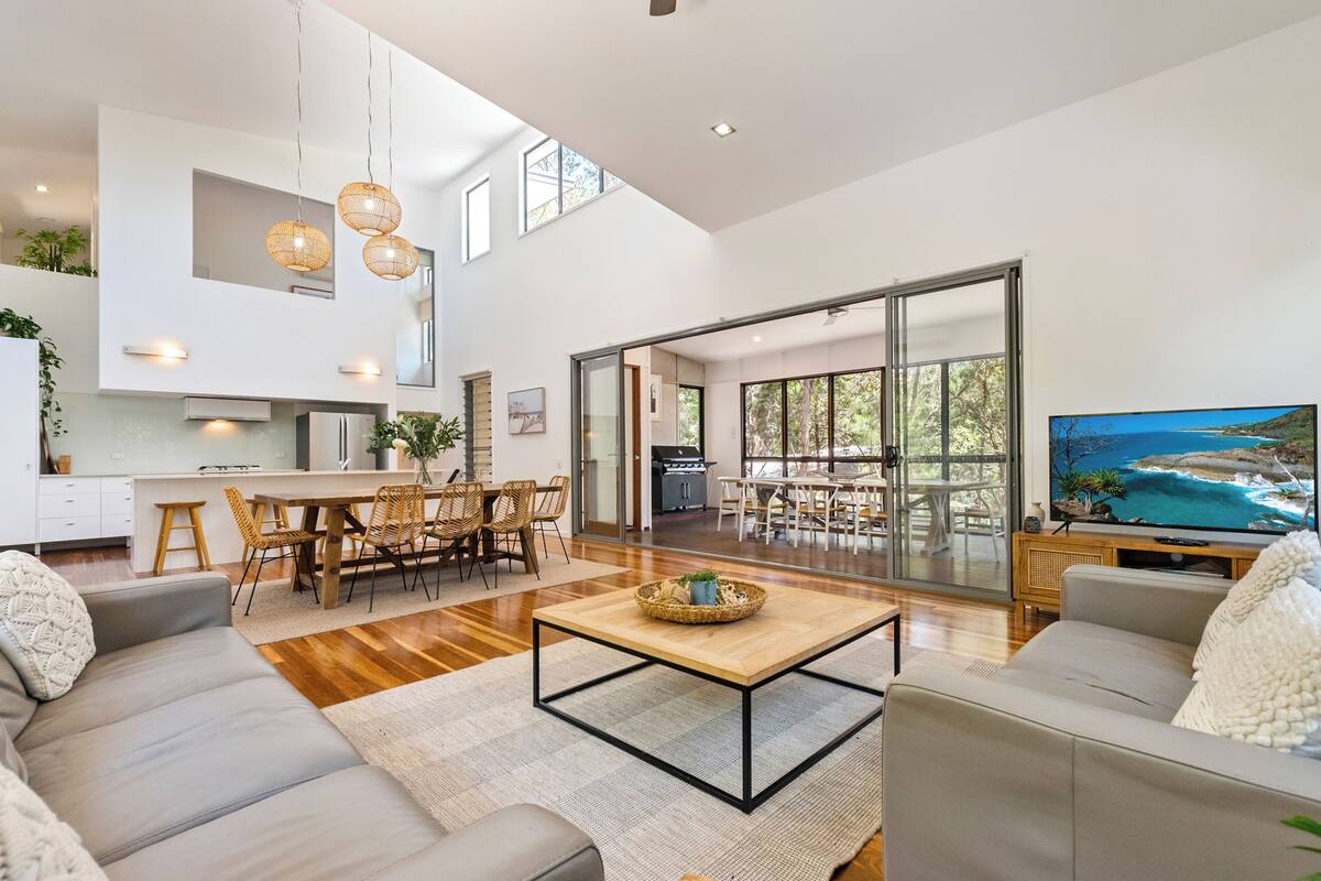 A spacious living area with a sofa, coffee table, and dining table. There is a TV on a wooden stand and a large window offering a view of the outdoors.