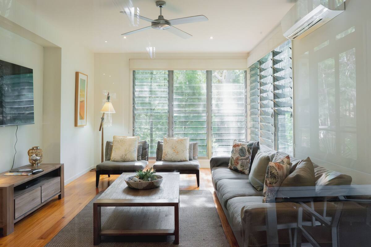 A spacious living room with a ceiling fan, a flat-screen TV, a coffee table with decorative items, two sofas with cushions, and large windows with blinds.