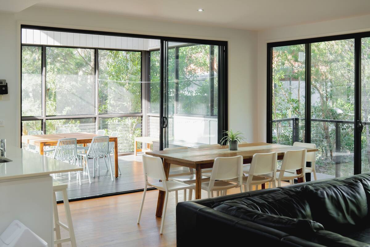 A modern living room with a wooden dining table, white chairs, a black leather sofa, and large windows offering a view of greenery.