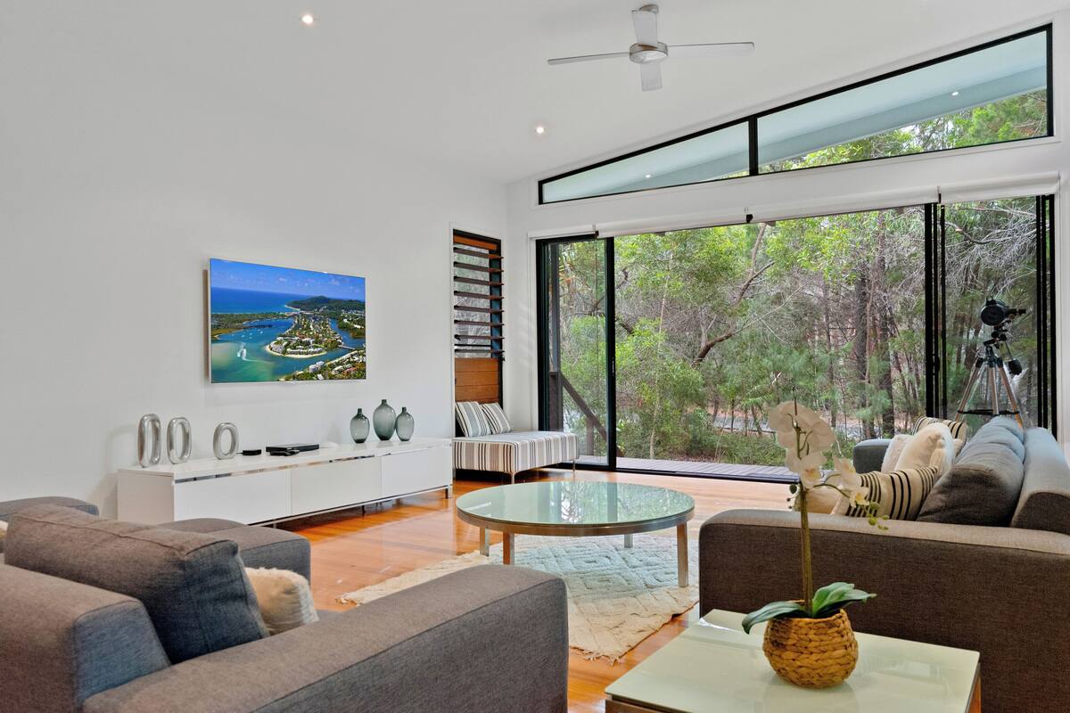 A modern living room with a large window, a round glass coffee table, and a painting of a coastal landscape.
