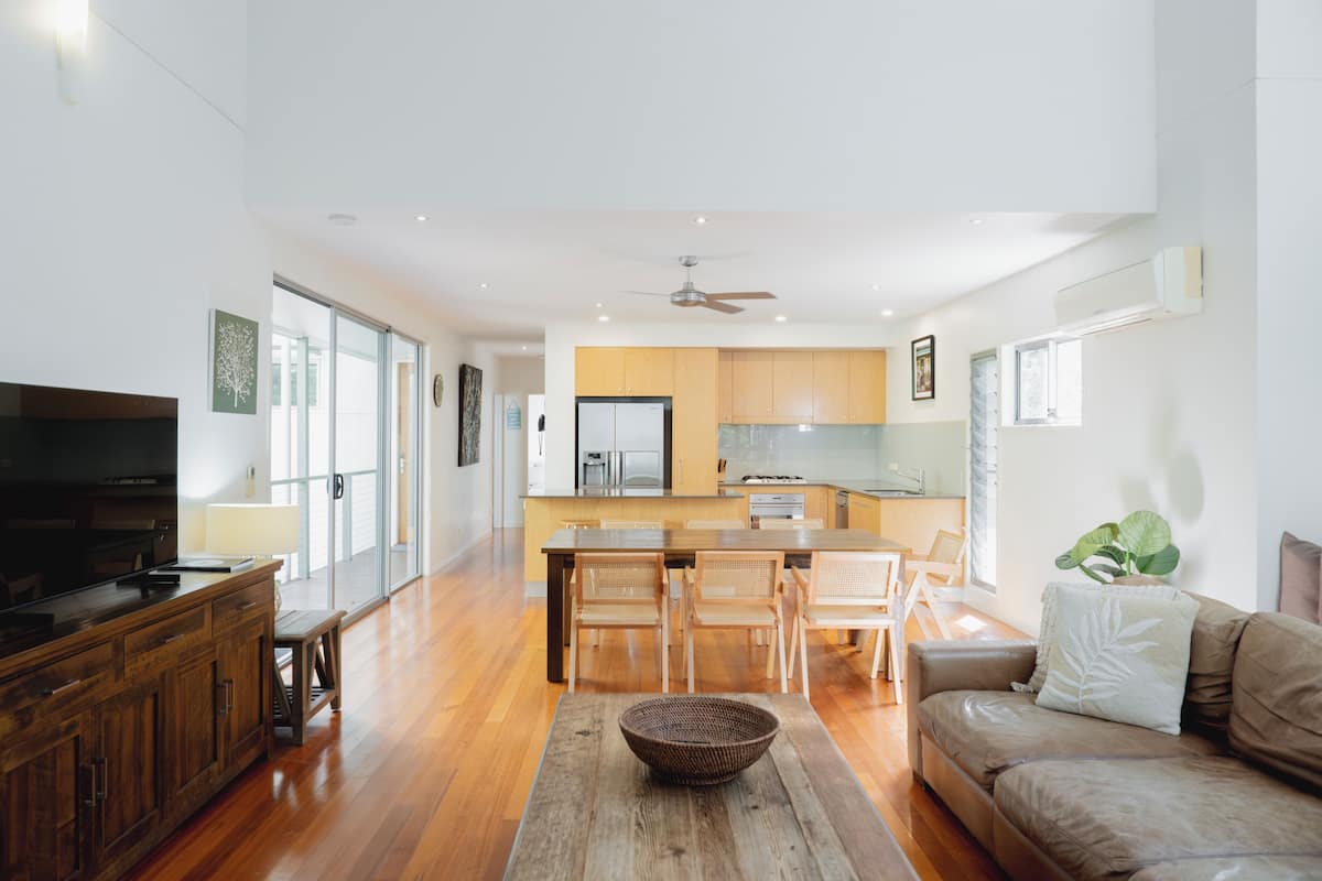 A modern living room with a wooden coffee table, a sofa, and a TV. There is a kitchen in the background with wooden cabinets and a dining area with chairs.