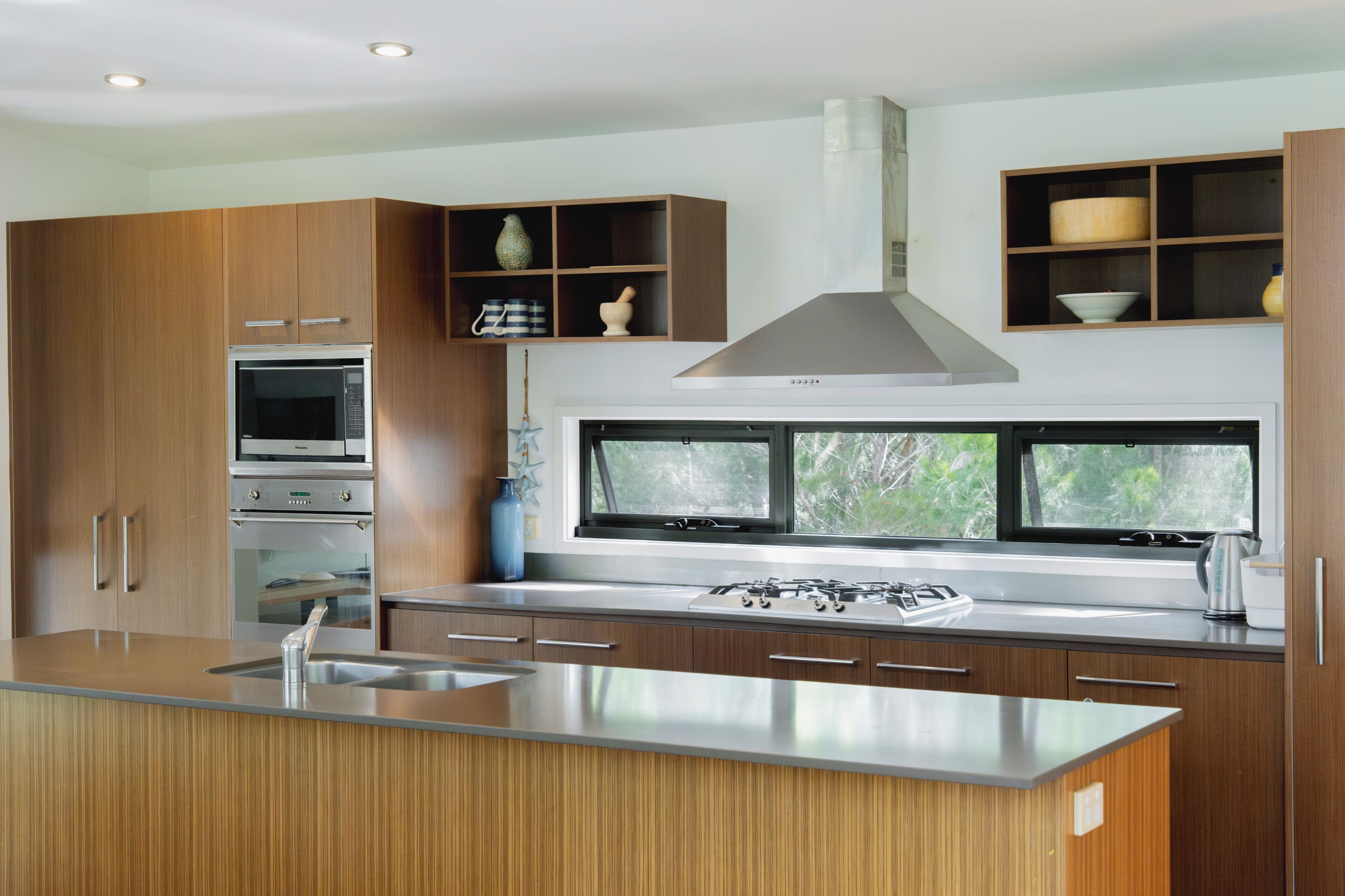Een moderne keuken met houten kastjes, roestvrijstalen apparatuur en een groot raam met uitzicht op groen.