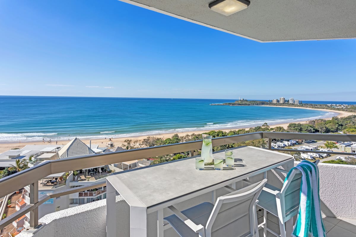 A balcony with a view of the beach and ocean, a table with glasses, and a towel.