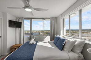 A hotel room with a bed, a ceiling fan, a TV, and large windows offering a city view.
