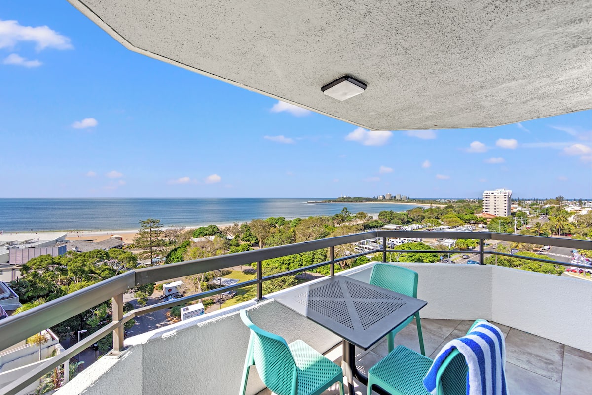 A balcony with a table and chairs, overlooking a beach and ocean.
