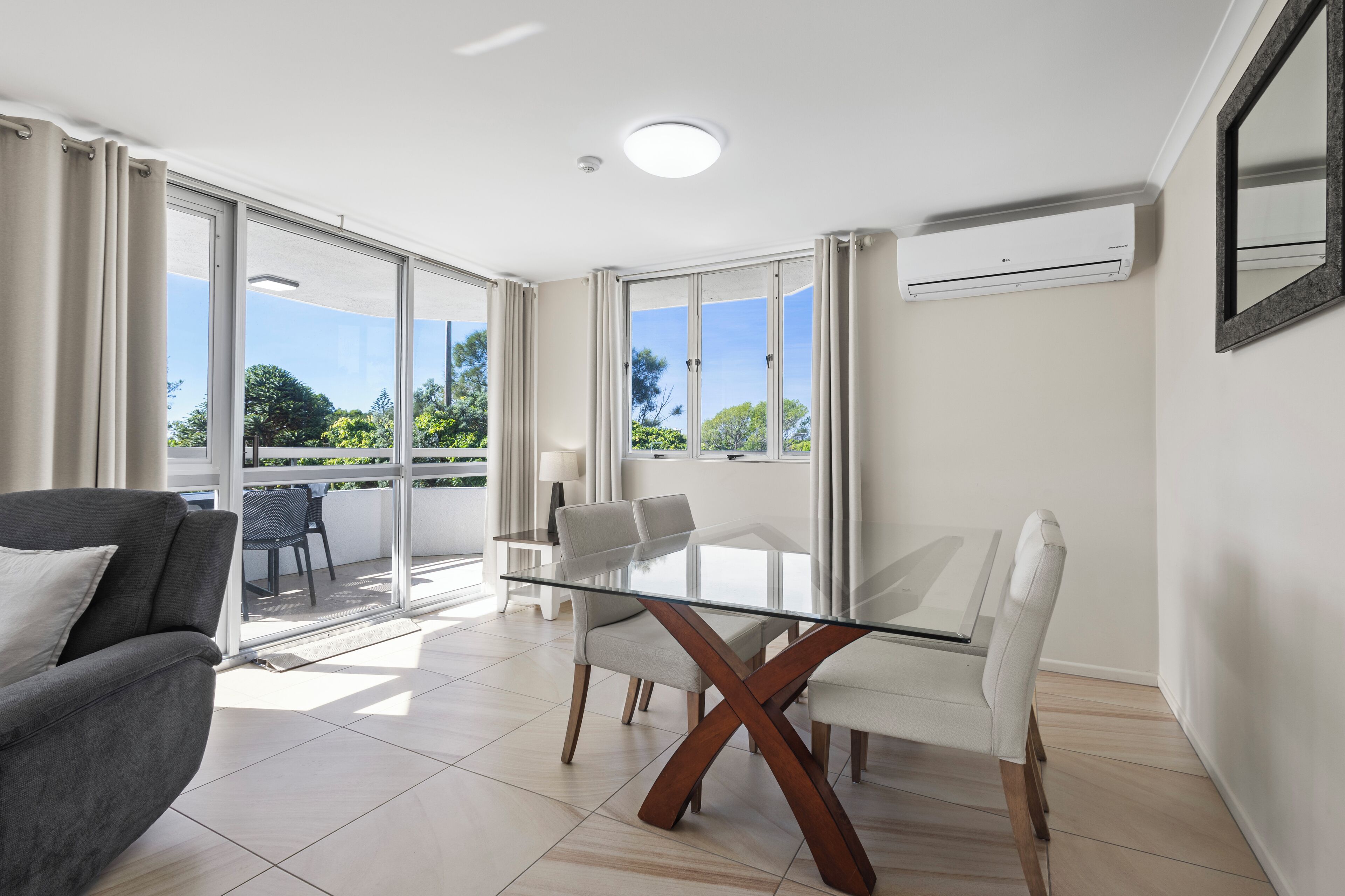 A modern living room with a glass-top dining table, white chairs, a grey sofa, and a balcony with a view of greenery.