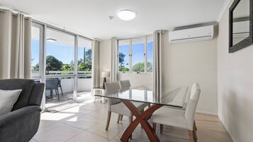 A modern living room with a glass-top dining table, white chairs, a grey sofa, and a balcony with a view of greenery.