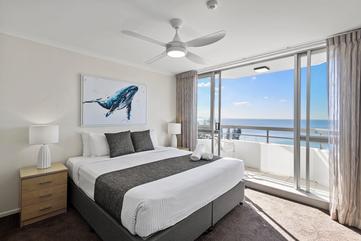 A modern hotel room with a large bed, a ceiling fan, a view of the sea, and a balcony with curtains.