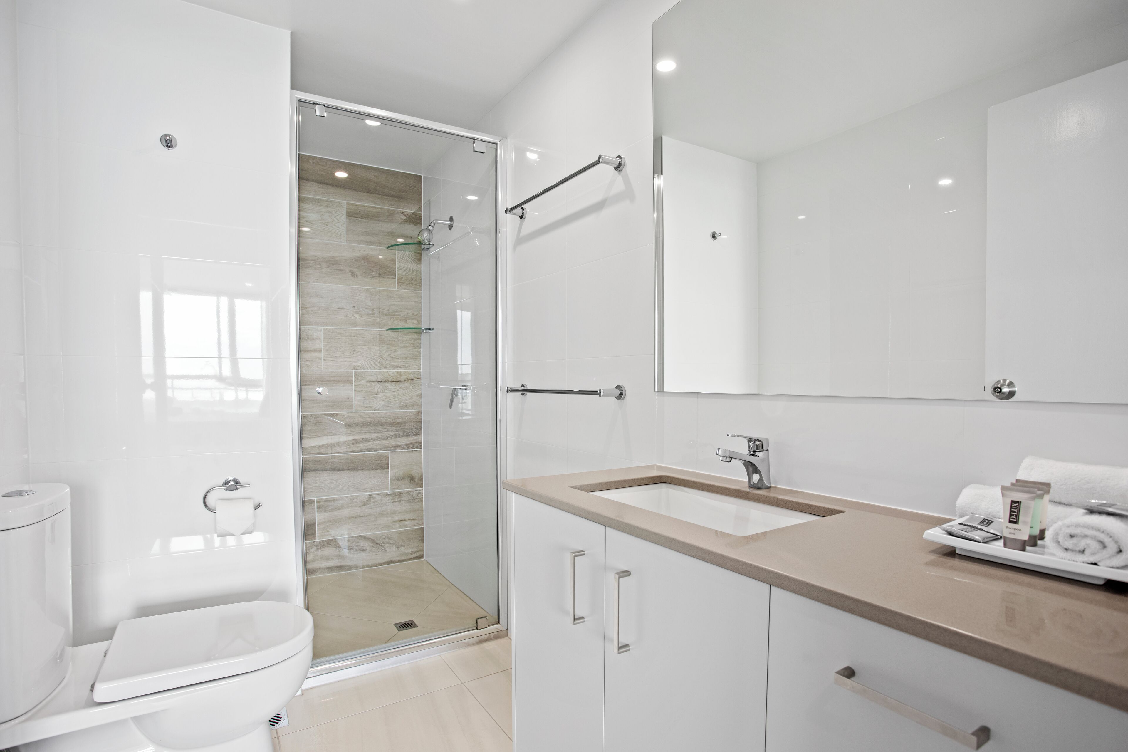 A modern bathroom with a glass shower enclosure, a white toilet, a beige vanity with a sink, and a large mirror.
