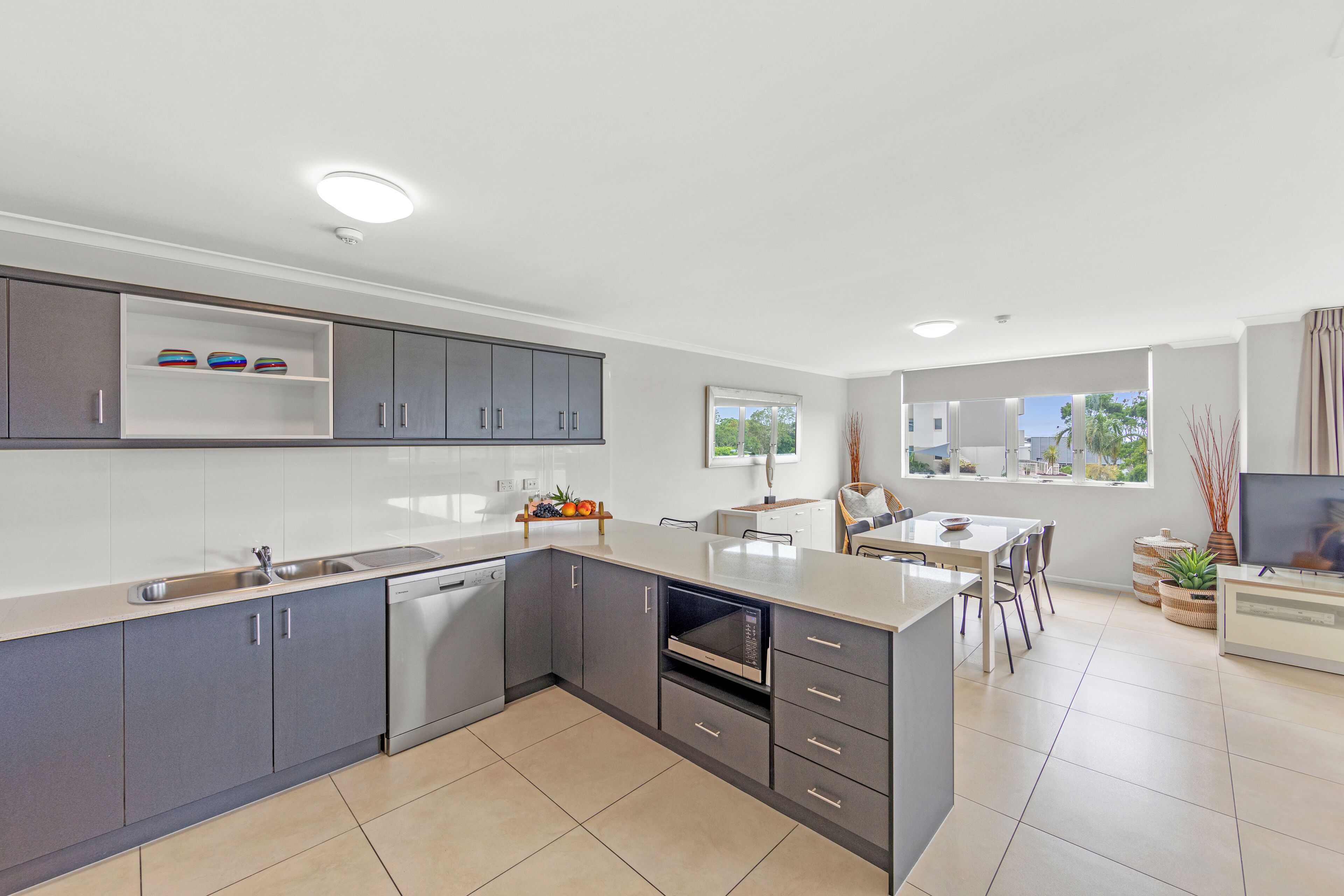 A modern kitchen with grey cabinets, a dishwasher, a built-in oven, and a dining area with a table and chairs.