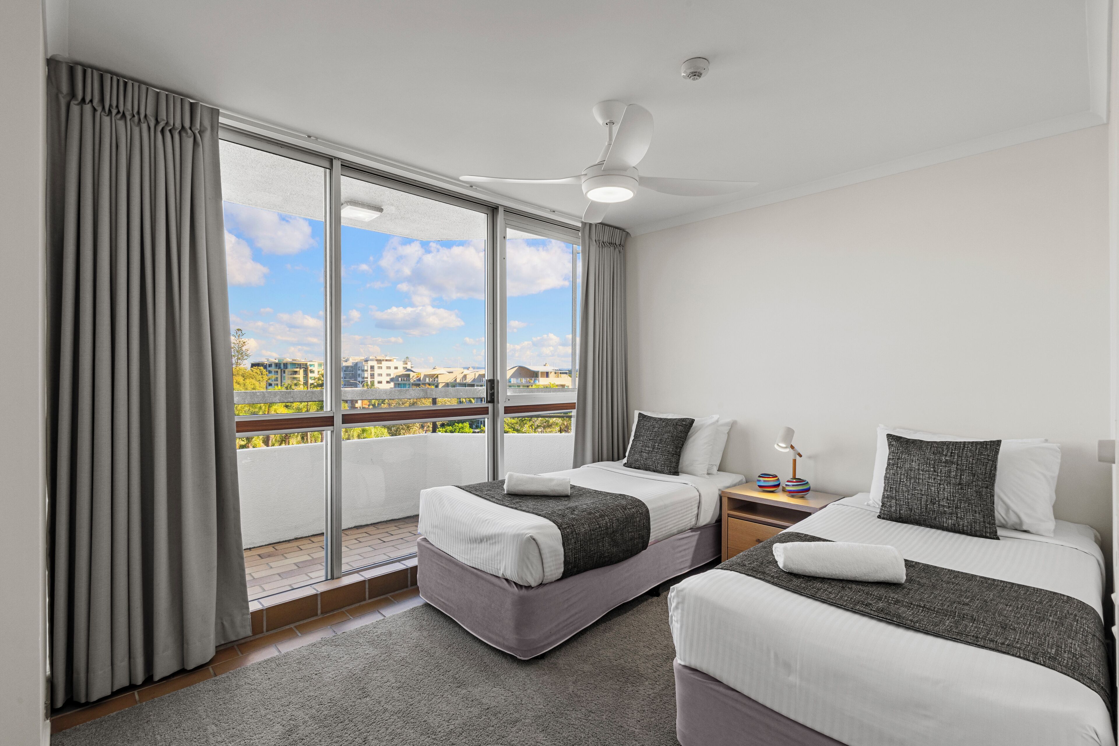 Ein Hotelzimmer mit zwei Betten, einem Balkon und Blick, einem Deckenventilator und einem Nachttisch mit Lampe.