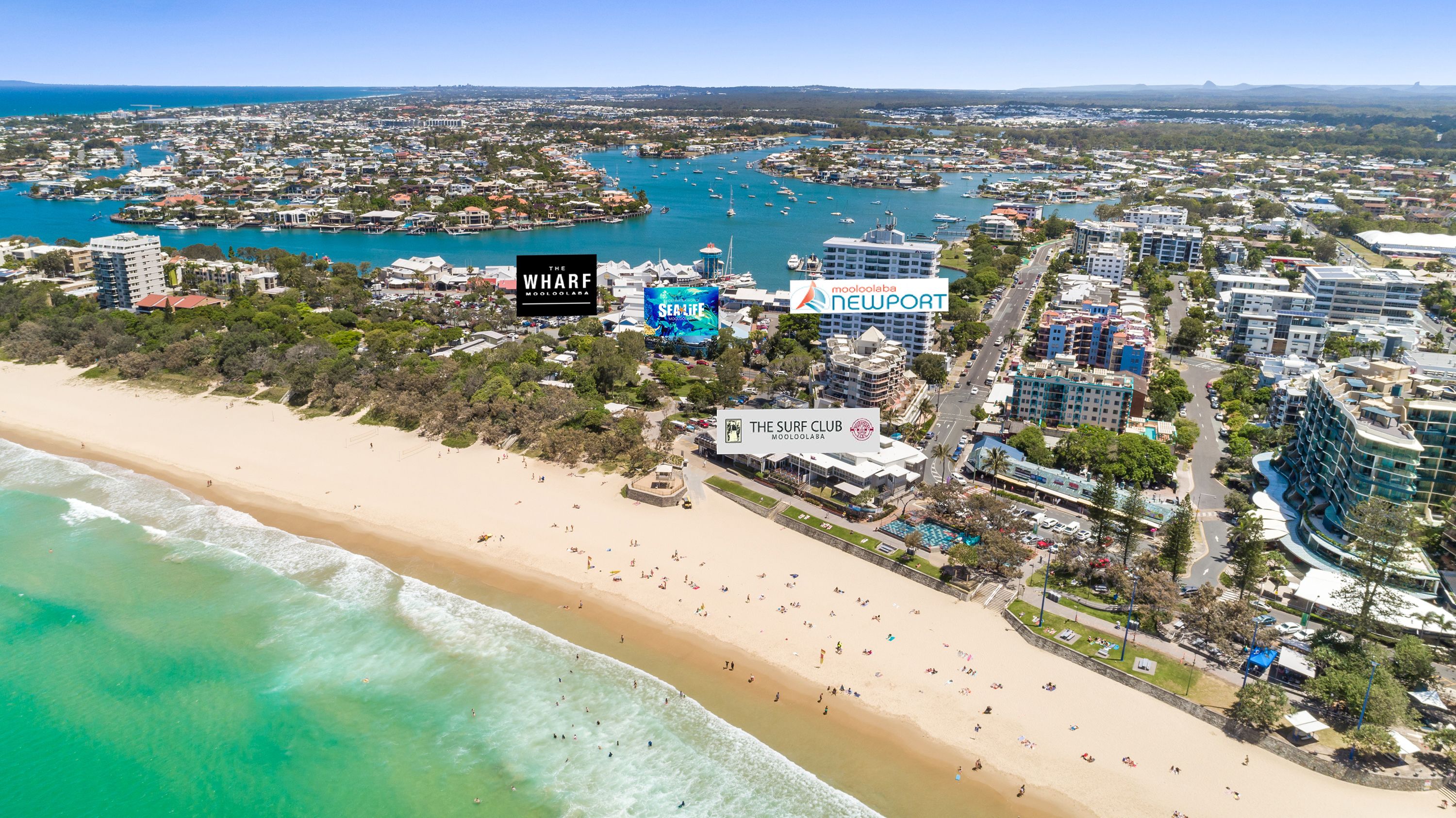 A coastal cityscape with a sandy beach, marina, and various buildings including hotels and a surf club.