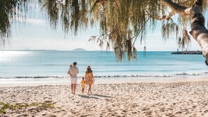 A sandy beach with a family walking along the shore, a clear sky, and a distant view of a rocky outcrop.