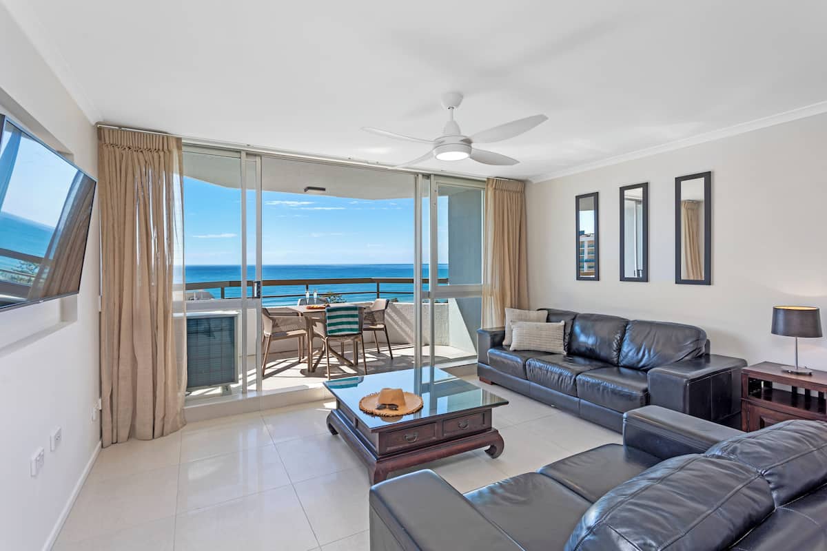 A spacious living room with a large glass door leading to a balcony with a view of the ocean. There is a ceiling fan, a sofa, a coffee table, and a TV mounted on the wall.