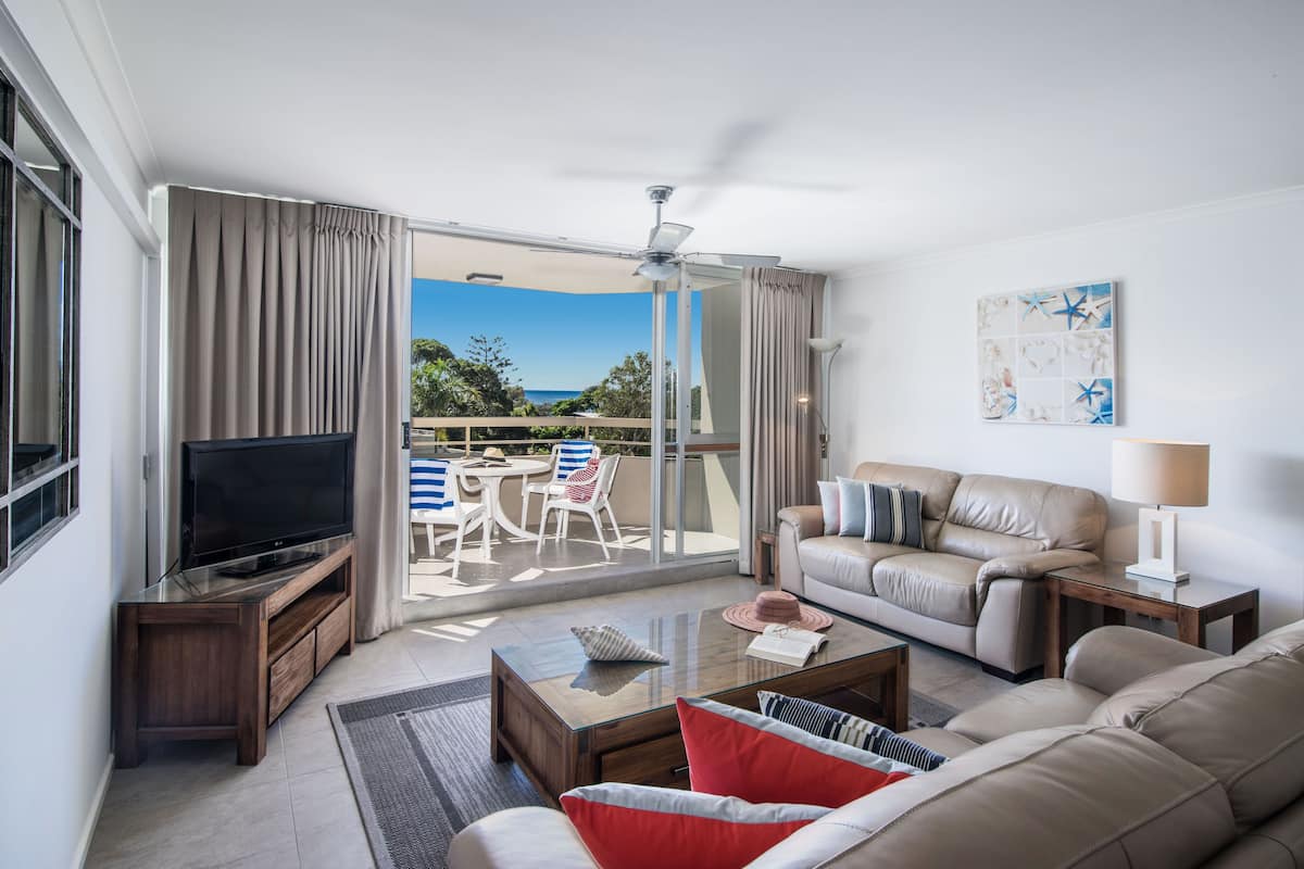 A modern living room with a flat-screen TV, leather sofa, and a glass coffee table. There is a balcony with a view of the ocean and a ceiling fan.