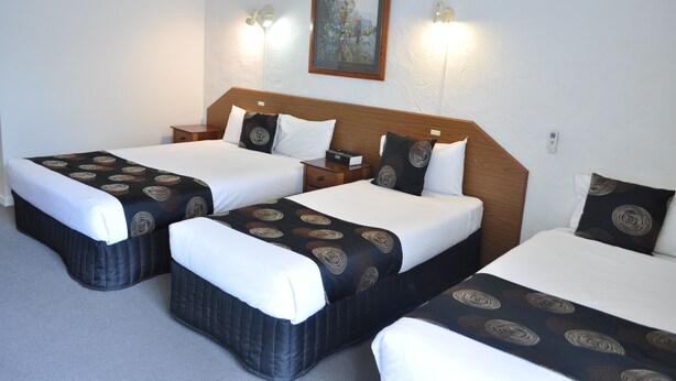 A hotel room with three beds, each with a patterned bedspread and pillows, a wooden headboard, bedside tables, and wall-mounted lights. - Albury Burvale Motor Inn (Albury)