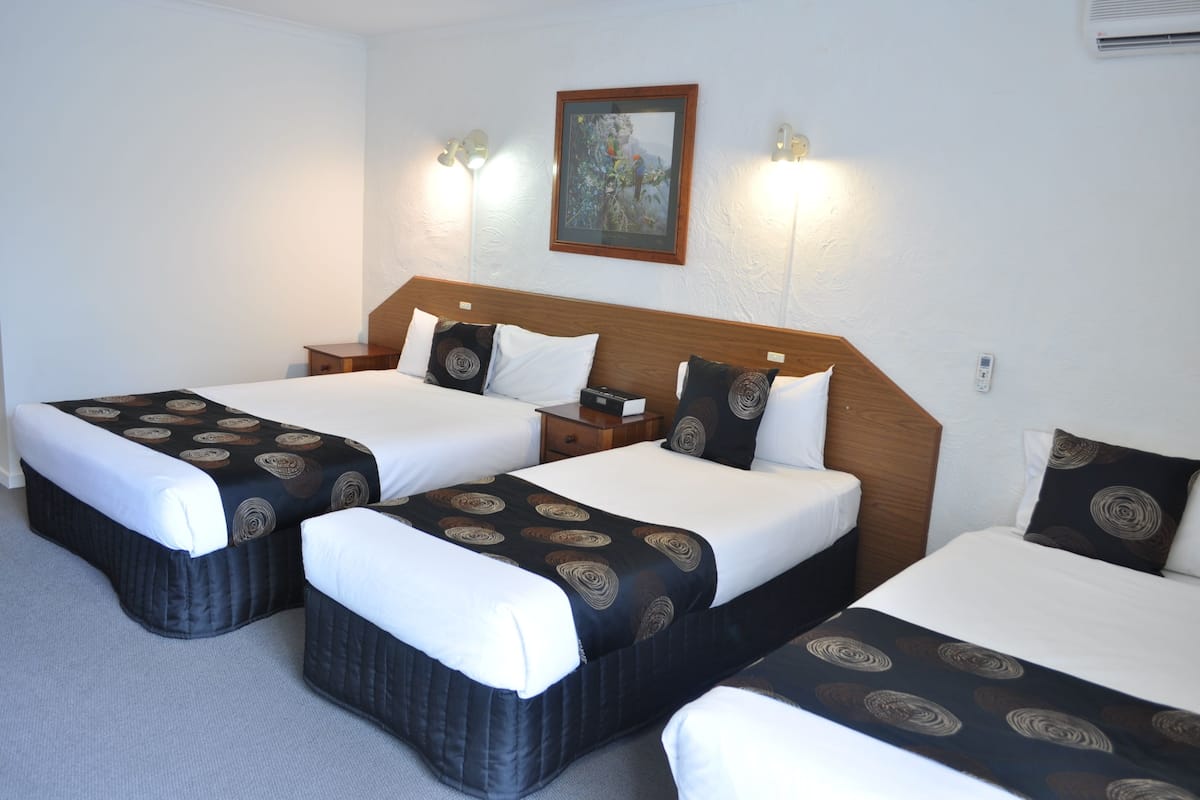Ein Hotelzimmer mit drei Betten, jeweils mit gemusterten Bettdecken und Kissen, einem Holz-Kopfteil, Nachttischen und Wandleuchten.