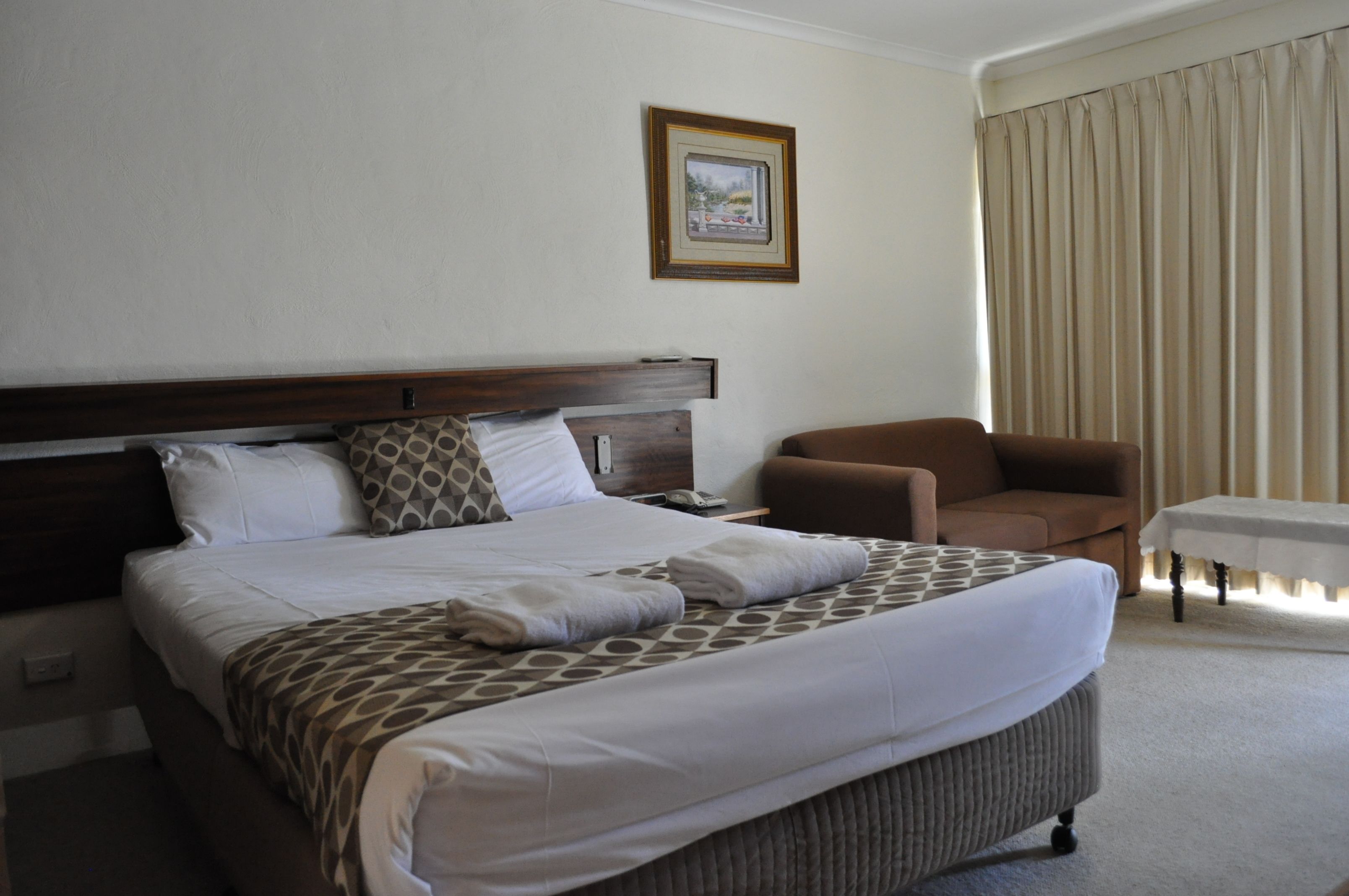 A hotel room with a large bed, a brown armchair, a small table, and a framed picture on the wall.