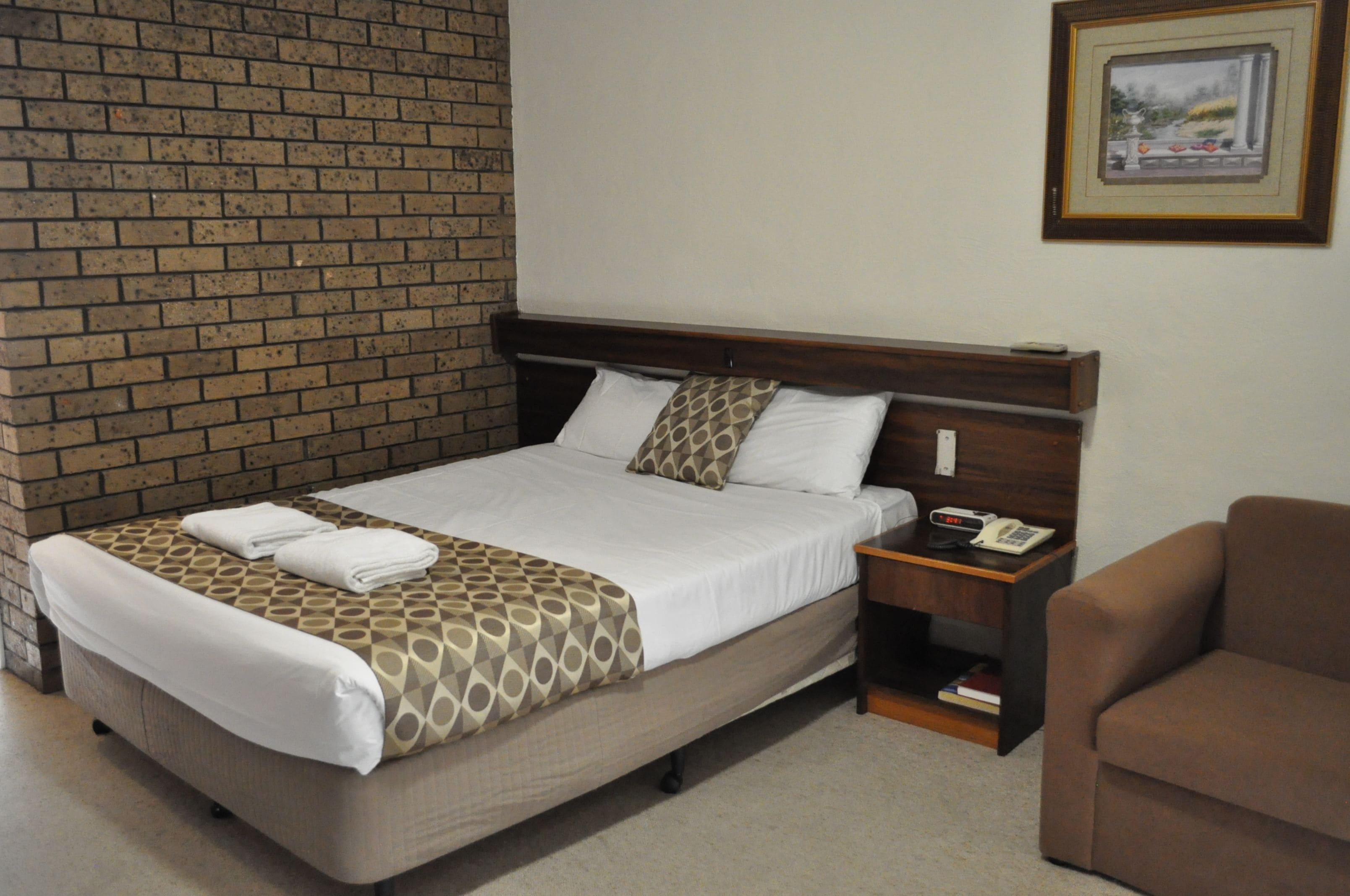 A hotel room with a brick wall, a bed with a patterned bedspread, a bedside table with a phone, and a brown armchair.
