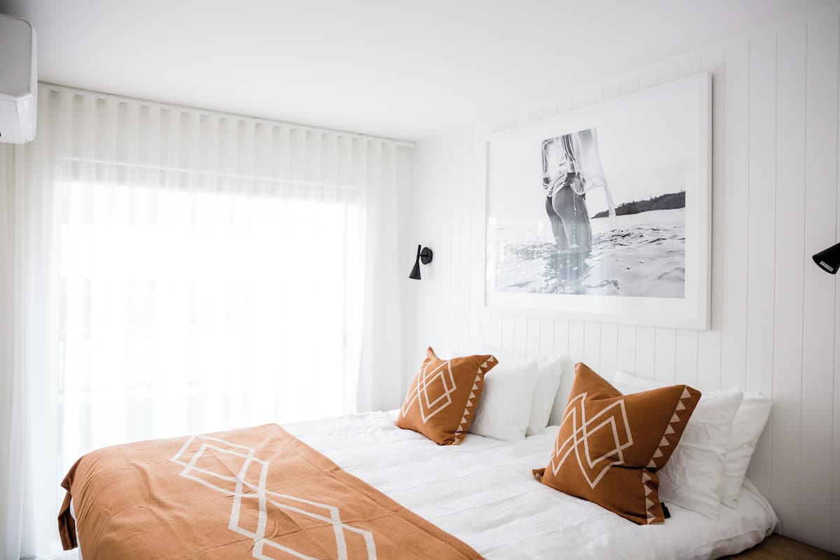Una cama bien hecha con un plaid de estampado geométrico, ropa de cama blanca y dos almohadas decorativas. En la pared, encima de la cama, se exhibe una fotografía blanco y negro.
