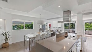 A modern kitchen with a central island, dining area with chairs, and large windows offering a view of greenery. - Saks on Hastings (Noosa Heads)