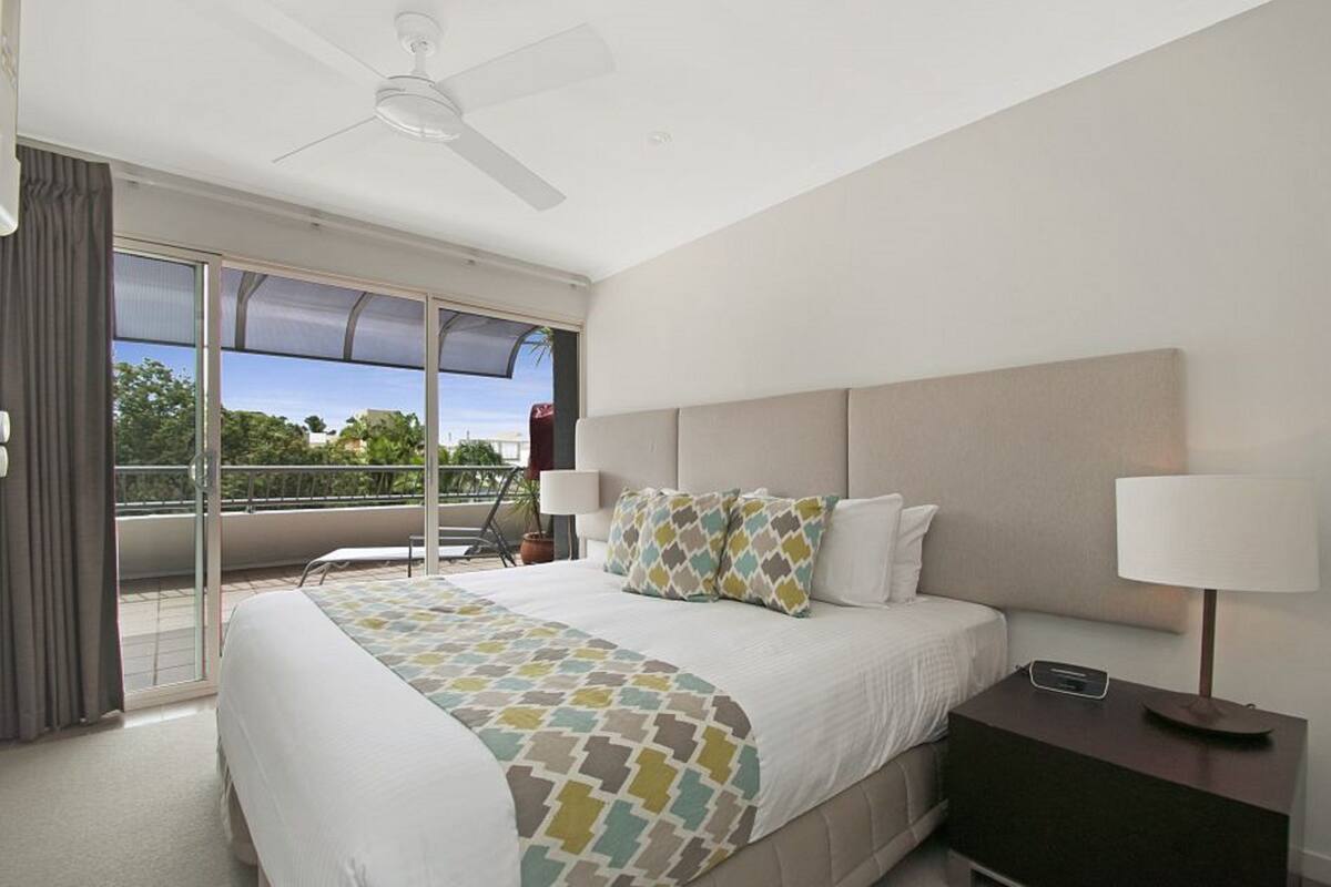 Habitación de hotel con una cama grande, ventilador de techo, balcón con vistas y mesita de noche con lámpara.
