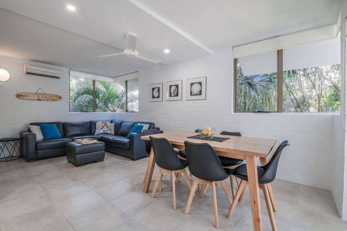 Una sala de estar moderna con sofá, mesa de comedor y sillas. Amplias ventanas con vistas al exterior, lleno de vegetación.