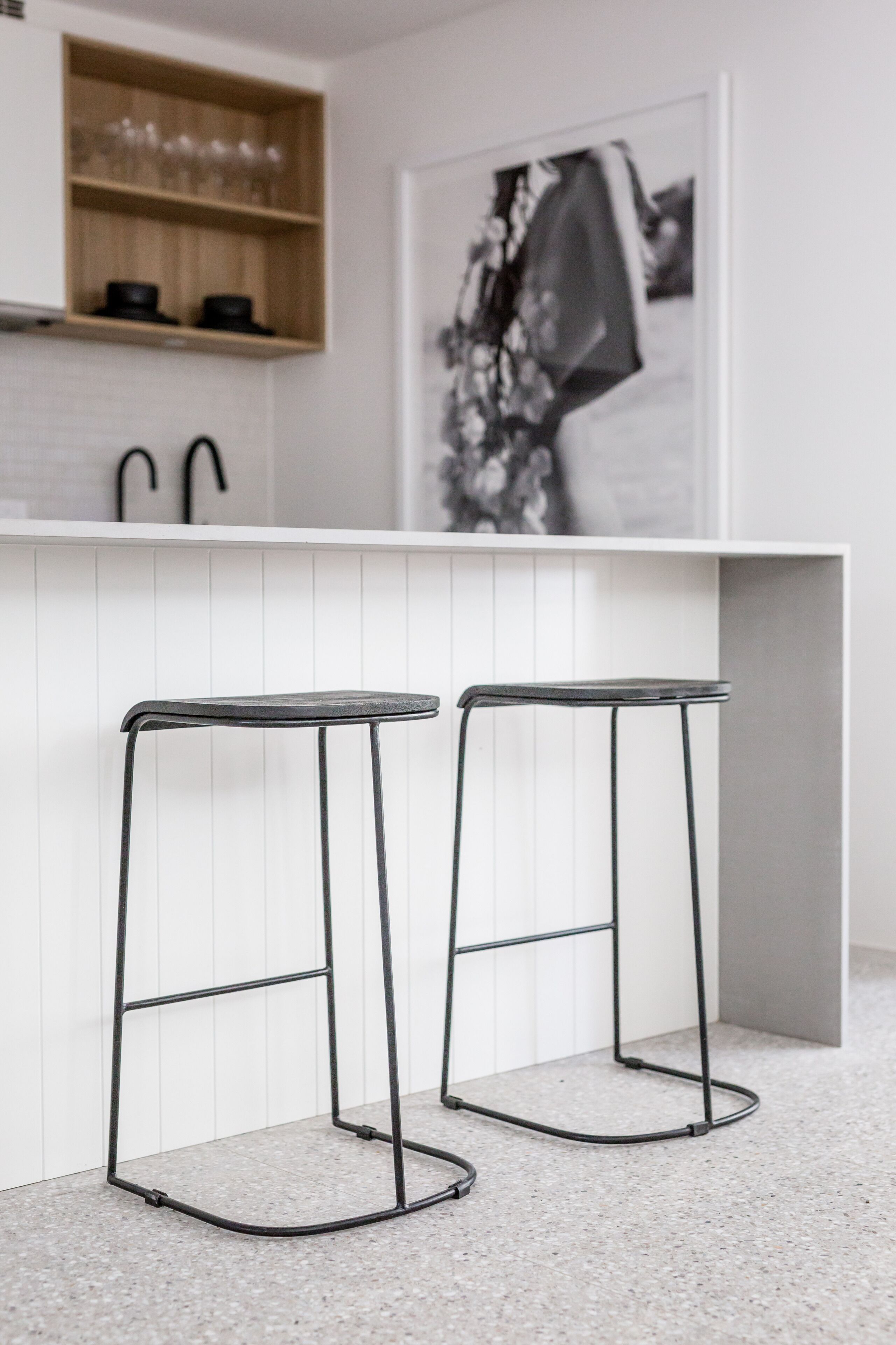 A modern kitchen with two black metal bar stools, a white countertop, and a wooden shelf above the sink.
