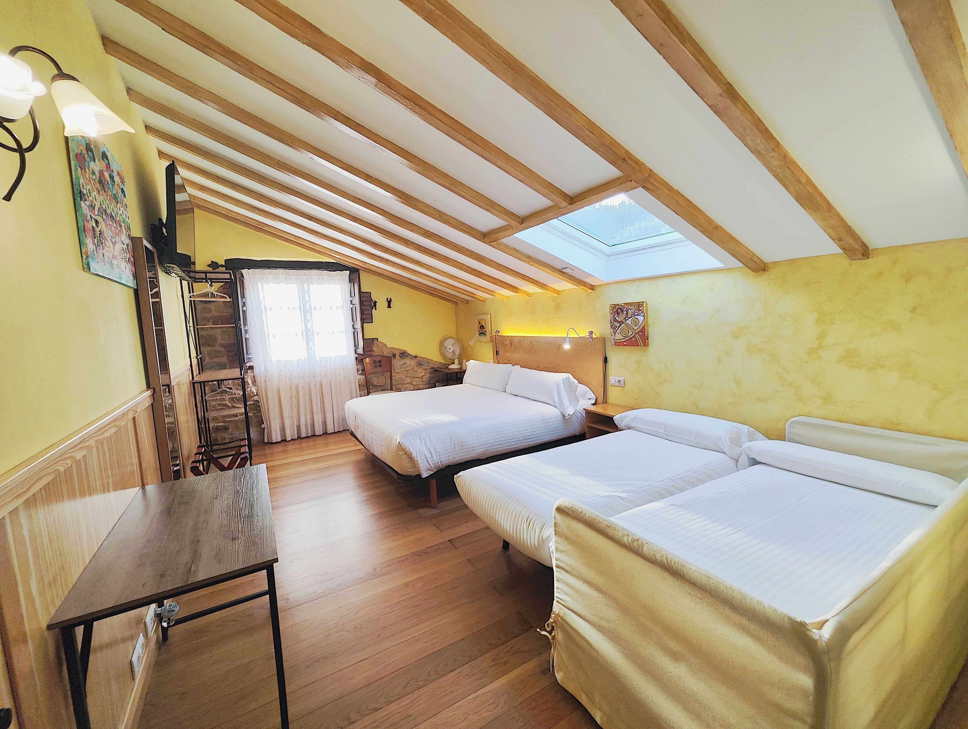 A cozy room with wooden beams, a skylight, a bed, a table, and a staircase leading to a balcony.