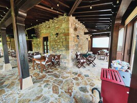 Un patio con suelo de piedra, mesas y sillas de madera, paredes de piedra y un cesto de basura rojo.