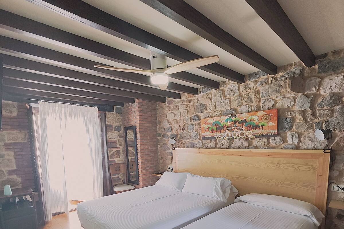 Une chambre avec deux lits simples, un plancher en bois, un mur en pierre et un ventilateur de plafond.