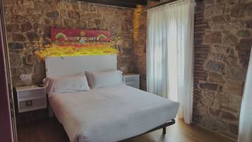 A bedroom with a stone wall, a bed with white bedding, a nightstand, a bedside table, and a window with curtains.