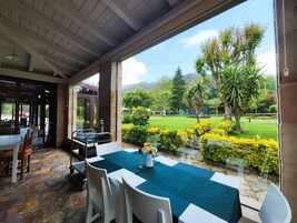 Un área de comedor al aire libre con una mesa preparada para dos personas, cubierta con un mantel verde, y vistas a un jardín bien cuidado con diversas plantas y césped.