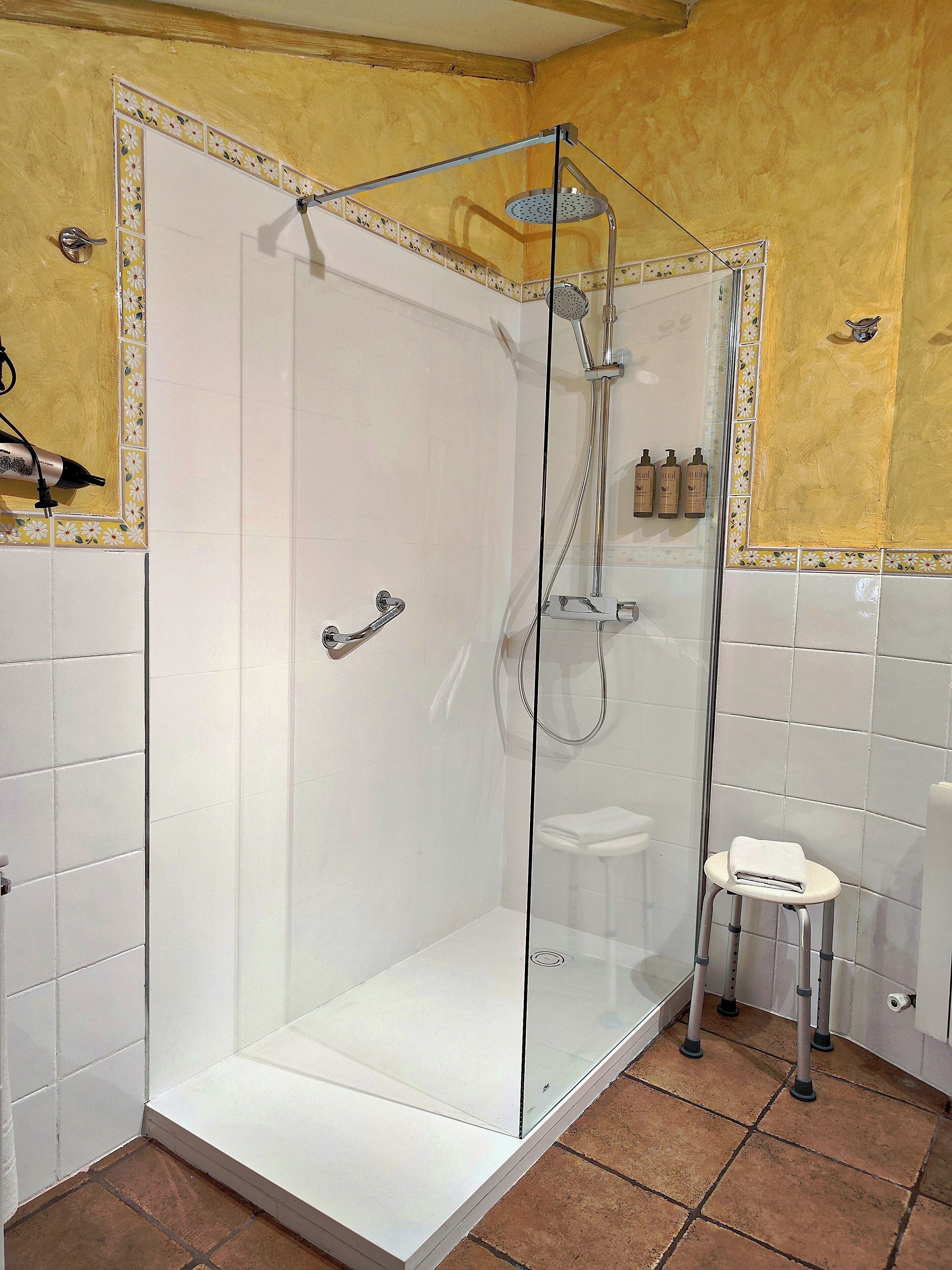 A glass-enclosed shower with a white tiled floor, a small stool, and a shelf with toiletries.